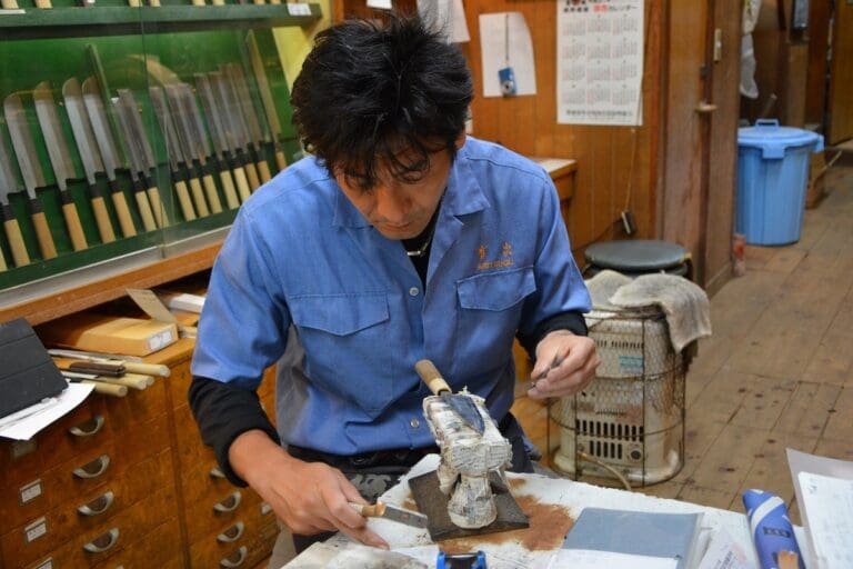 How to Make Your Own Knife in Japan: Best Classes Guide Japanese man in blue workshop jacket sharpening a knife in a knife workshop