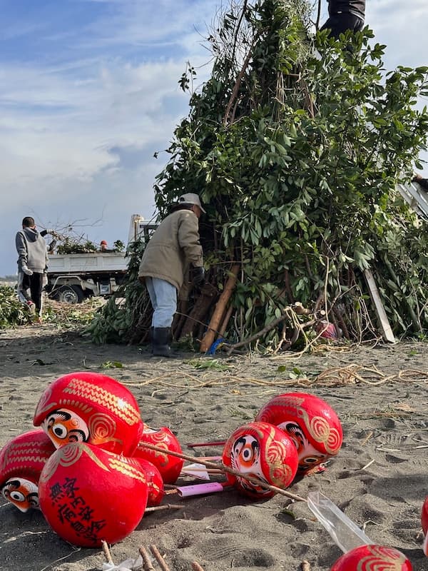 A straw structure being prepared for the Oiso Sagicho Festival, with Daruma dolls placed nearby and people attaching greenery to the frame.