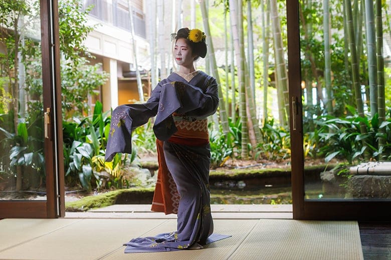 Maiko performing a traditional dance indoors at Fortune Garden restaurant in Kyoto, with a bamboo garden visible through the open doors