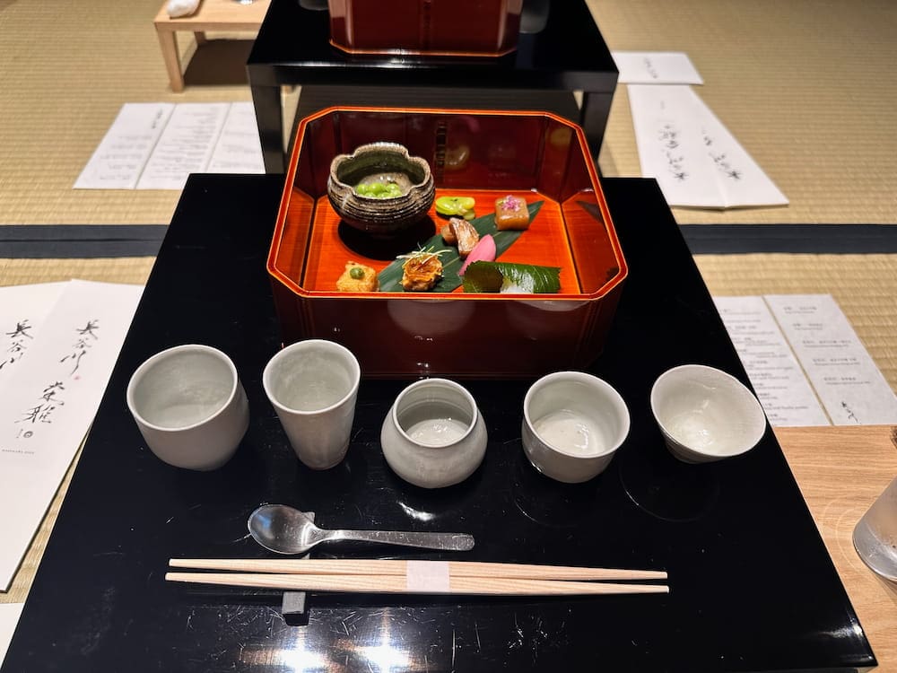 Close-up of sake tasting setup with five cups and a lacquered food tray containing seasonal dishes.