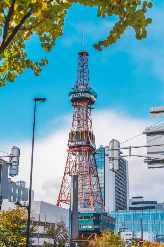 Red Sapporo TV Tower with Panasonic digital clock rising above city buildings and autumn trees under a blue sky