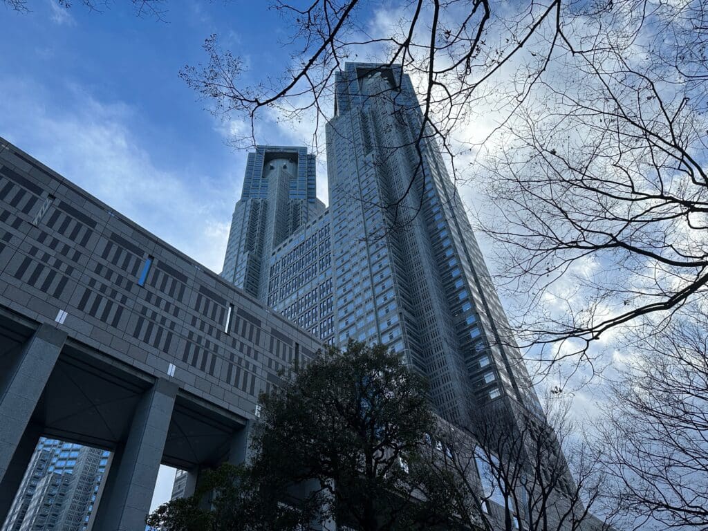 Low-angle view of Tokyo Metropolitan Government Building twin towers framed by bare tree branches under a cloudy blue sky