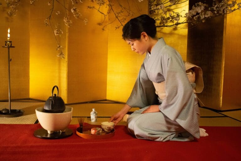Woman in kimono performing Japanese tea ceremony with kettle and utensils on tatami before a gold screen and cherry blossoms