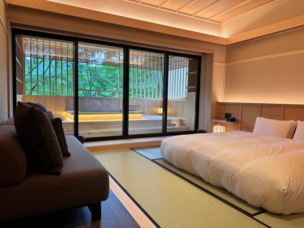 Hakone Nazuna Ryokan room with a low platform bed, tatami mats, and a sliding glass window looking onto a green forest garden
