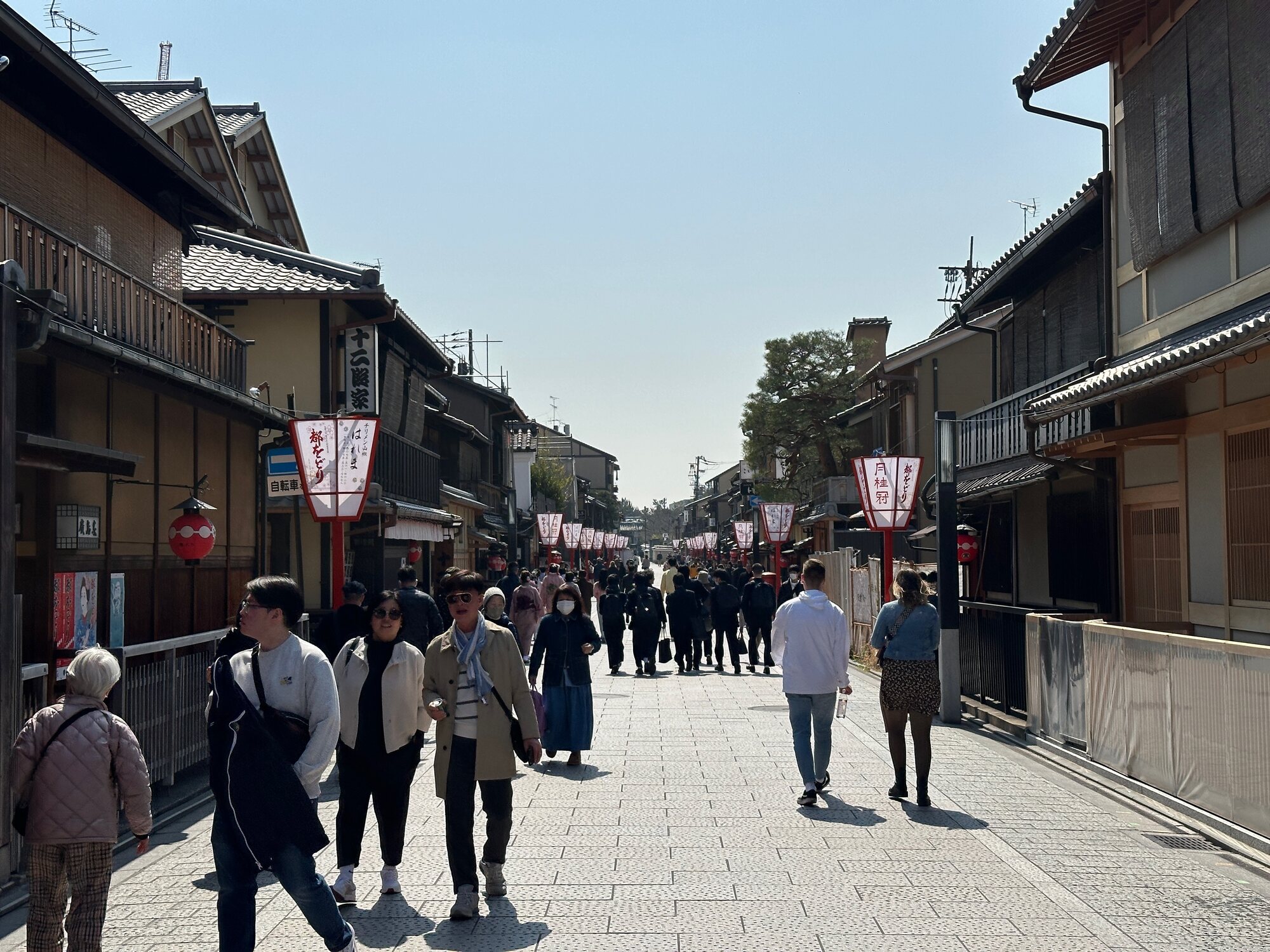 Crowds walking down a traditional street in Kyoto’s Gion district, lined with old wooden buildings and red lantern banners