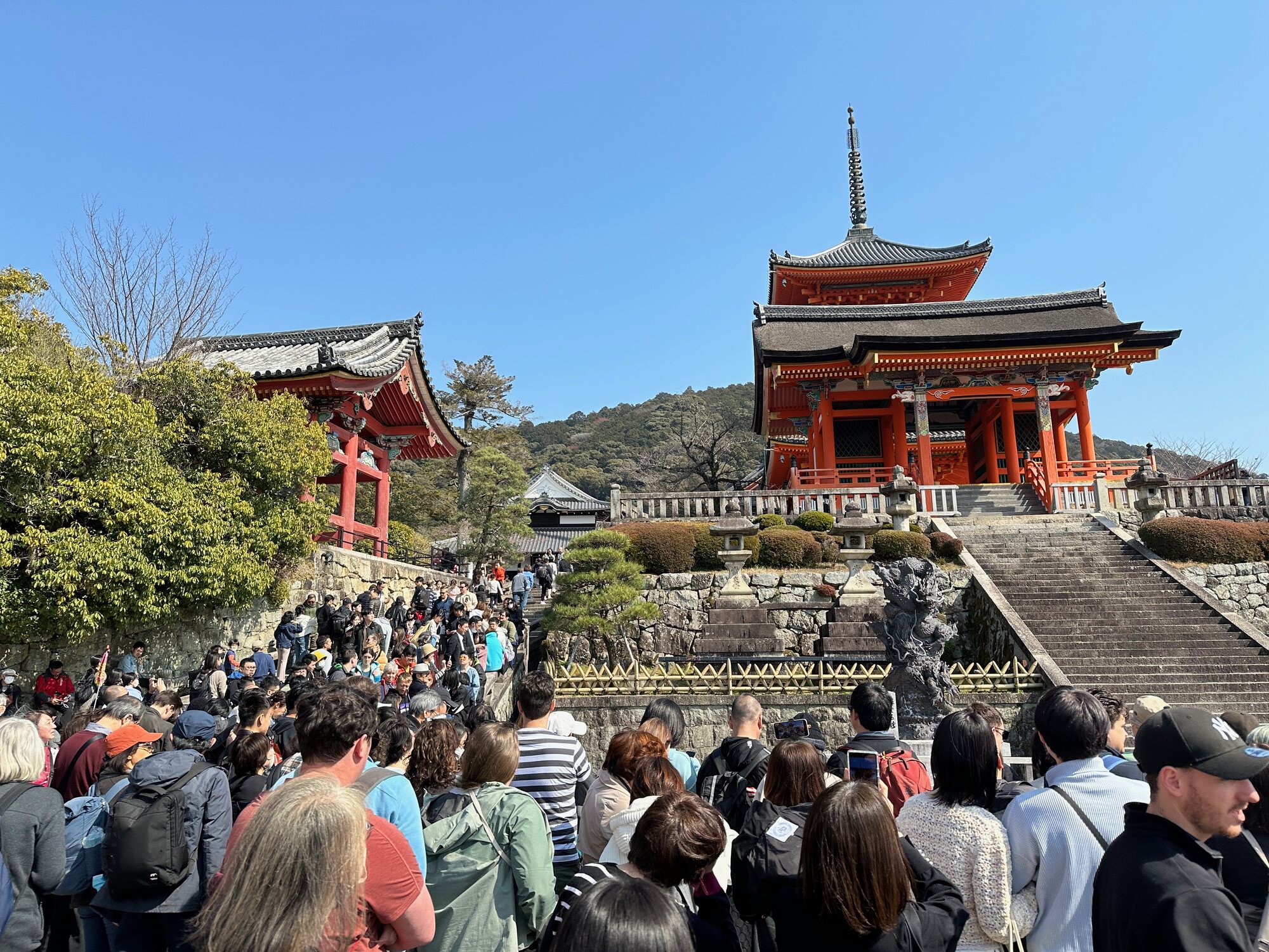 Crowd walking up the stone steps toward Kiyomizu-dera in Kyoto, Japan, with red temple halls and a clear blue sky