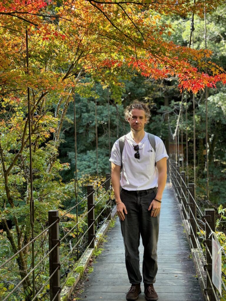 Man standing on a narrow wooden walkway through an autumn forest with red and yellow maple leaves overhead