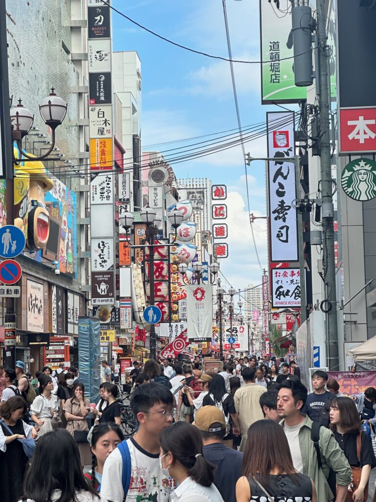 Crowded shopping street in Osaka with many storefront signs and banners, including a large vertical sign and the Starbucks logo