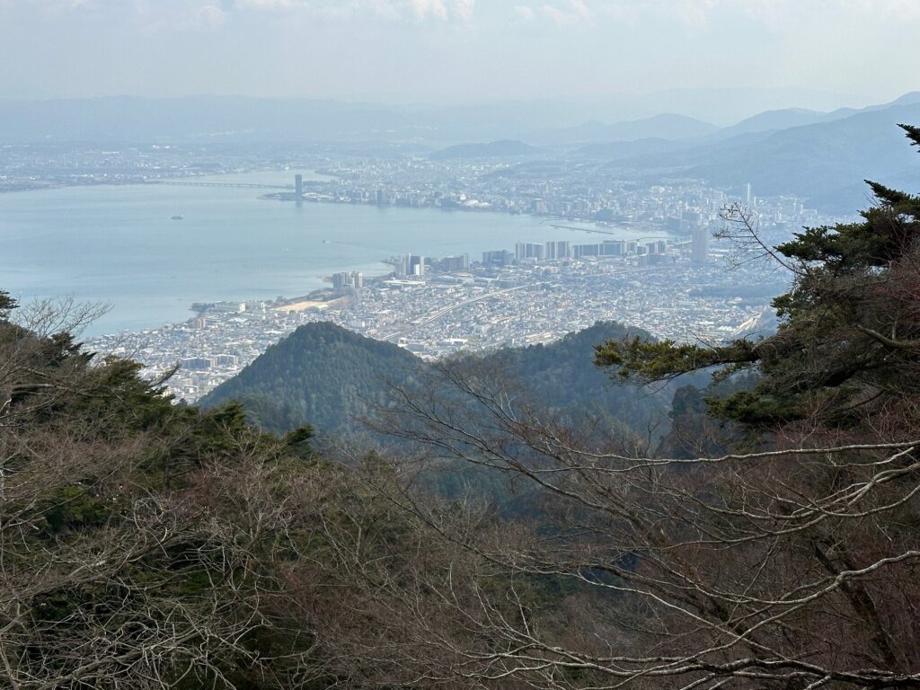 View over Lake Biwa and Otsu city from a mountainside lookout, with pine trees in the foreground and a bridge crossing the lake