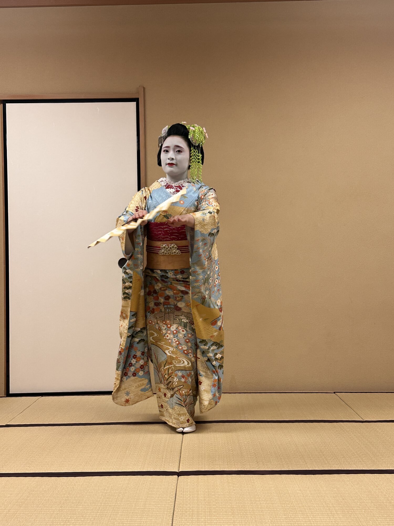 A geisha performer in colorful kimono holding a fan during a dance on a tatami stage in Tokyo